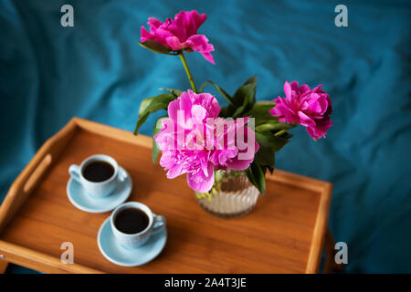 Belles pivoines rose dans un vase de verre et deux tasses de café sont debout sur un plateau en bois dans le lit. Banque D'Images