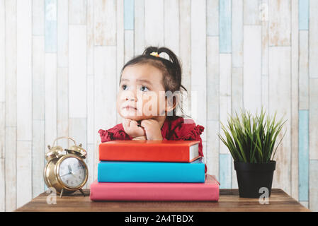 Cute little asian girl gazing et regardant l'espace vide contre livres sur table. Concept de l'éducation, l'avenir et la rêverie Banque D'Images