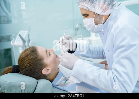 La prévention de la carie dentaire dentiste professionnel à l'aide d'instruments de restauration pour les dents primaires. Female patient lying on dentiste président. Le port de cap médical Stomatologist, gants et masque. Banque D'Images