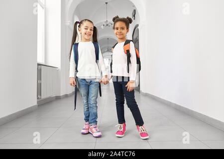 Deux écolières heureux avec sac à dos pour l'école derrière les épaules debout dans long couloir de l'école et tenir la main. Les élèves de l'école primaire positif et souriant à la caméra à. Banque D'Images