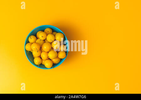 Modèle d'été créatif à base de produits frais dans les prunes cerises jaunes bol bleu sur fond jaune pastel. Concept minimal de fruits. Mise à plat. Banque D'Images