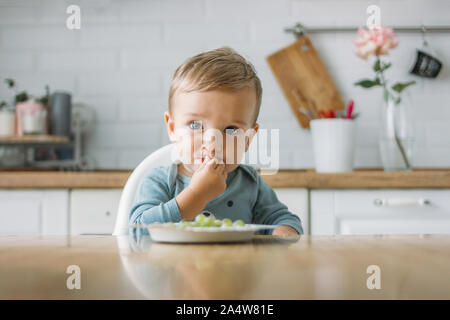 Charmant petit garçon peu concentré d'abord manger au raisin vert alimentaire cuisine lumineuse à la maison Banque D'Images