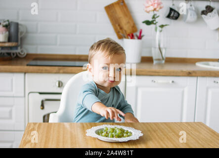 Charmant petit baby boy eating première nourriture à raisin vert la cuisine lumineuse à la maison Banque D'Images