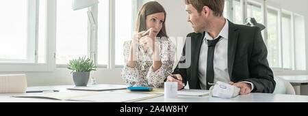 Deux collègues, un jeune homme et femme, dans une discussion approfondie sur les comptes qu'ils sont assis à un bureau à l'aide d'une table ajout manuel de la machine. Banque D'Images