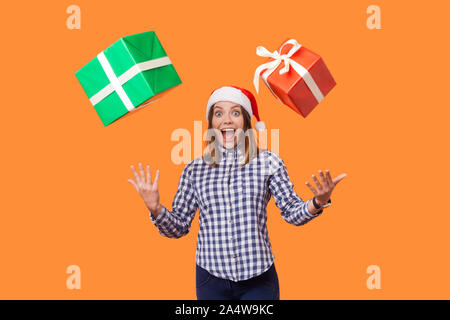 Portrait de stupéfié brunette woman in santa hat et chemise à carreaux debout avec la bouche ouverte et les mains levées, en essayant d'attraper le vol des cadeaux de Noël. Banque D'Images