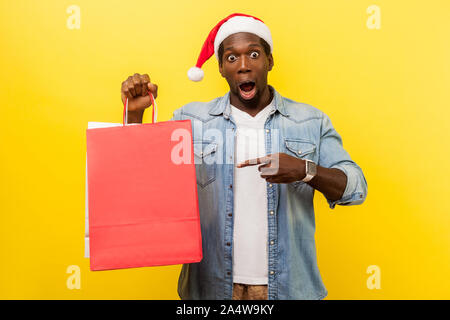 Portrait de surpris handsome young man in santa hat et occasionnels chemise en jean debout, pointant sur sac de shopping, à la recherche de rabais de noël choqué. Banque D'Images