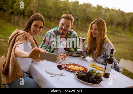 Groupe de jeunes gens assis près de la table, de boire du vin rouge et en tenant avec selfies téléphone mobile dans le vignoble Banque D'Images