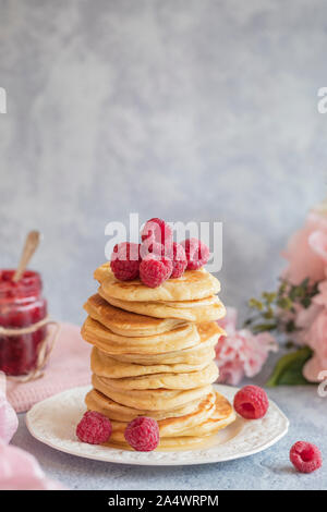 Une pile de crêpes américaines avec des framboises fraîches sur le dessus. Il y a un pot de confiture à l'arrière-plan, som fleurs et un tissu rose. L'arrière-plan est i Banque D'Images