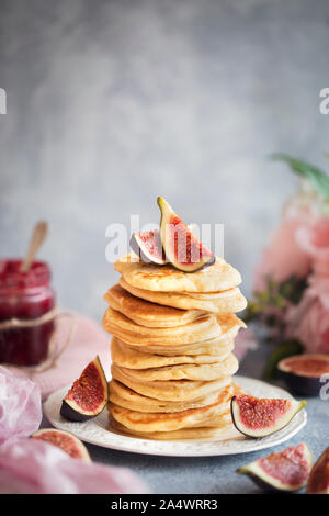 Un petit-déjeuner frais avec une pile de crêpes et de figues fraîches. Il y a un pot de confiture à l'arrière-plan, quelques fleurs, et un morceau de textile rose. Banque D'Images