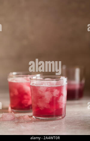 Un verre avec de la glace, rouge sur un fond gris. Il y a une glace broyée sur la table, et dans l'arrière-plan il y a deux verres defocused. Banque D'Images