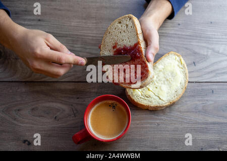 Une tranche de pain blanc, de confiture sur propagation il vus du dessus et une tasse à café rouge sur une table en bois Banque D'Images