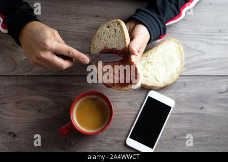 Une tranche de pain blanc, de confiture sur propagation il vus du dessus et une tasse à café rouge sur une table en bois Banque D'Images