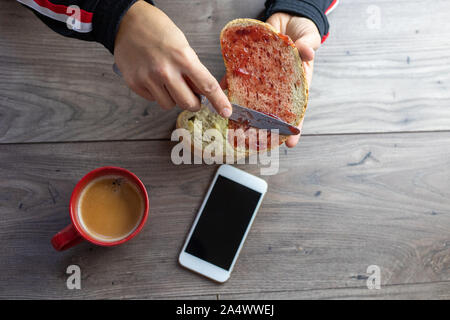 Une tranche de pain blanc, de confiture sur propagation il vus du dessus et une tasse à café rouge sur une table en bois Banque D'Images