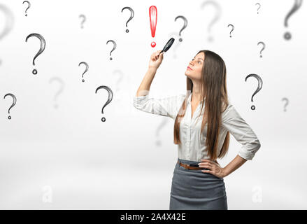 Young businesswoman with loupe sur sa tête à la recherche de point d'exclamation rouge entouré par des points d'interrogation Banque D'Images