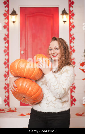 Cute adultes grosse fille en blanc chandail avec étoiles d'or détient une grande pile de citrouilles et se tient sur porche de maison blanche Banque D'Images