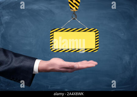 Un chèque en blanc à rayures jaune signe construction suspendue à un crochet de grue au-dessus de palm sur des hommes d'arrière-plan du tableau. Banque D'Images