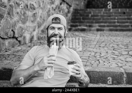 Ce qu'un casse-croûte. Homme barbu malsain de manger sandwich hot dog snack. Le repos et le Hipster ayant hotdog snack sur escalier extérieur. Guy caucasienne bénéficiant d'un casse-croûte sur rue. Banque D'Images