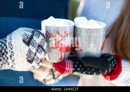 Close-up of a couple holding tasses avec cerfs avec une boisson chaude et des guimauves dans leurs mains dans des gants d'hiver. Banque D'Images