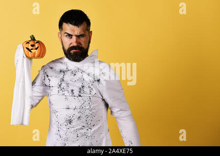 L'homme avec toute l'expression visage sur fond jaune, copiez l'espace. Heureux temps des Fêtes Halloween concept. Guy avec barbe est titulaire d'orange citrouille avec sourire. Caractère dans Halloween Costume fantôme blanc Banque D'Images