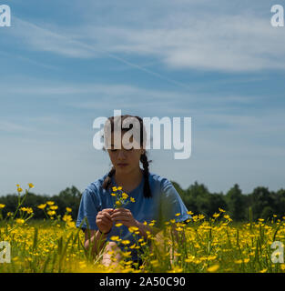 Teen girl holding Flowers et à les regarder alors qu'il était assis dans un grand champ avec des fleurs jaunes et des arbres en arrière-plan Banque D'Images