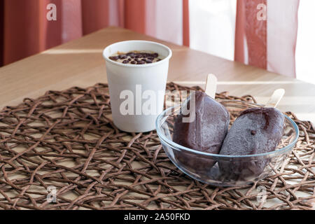 Végétalien premières glaces faites maison. Petite partie dans du papier blanc tasse et deux sucettes glacées sur un bâton. Fait à partir de la banane et la crème de coco, couverts de Banque D'Images