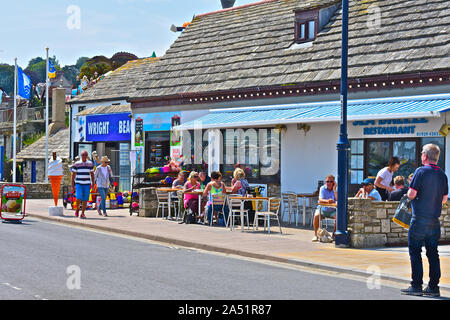 Maison de vacances typique de la scène avec des gens assis à l'extérieur d'un café tout en admirant le soleil d'été donnant sur la mer. Les vacanciers en passant devant. Banque D'Images