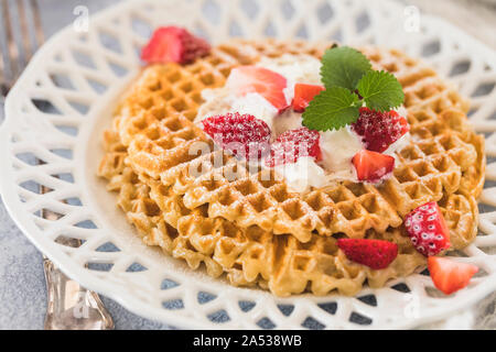 Gaufres d'or frais avec des morceaux de fraises et crème fouettée sur le dessus. Plaque à motifs blancs sur fond gris. Banque D'Images