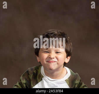 Studio shot horizontale d'un petit garçon faisant un visage stupide par scrunching son visage. Fond brun avec copie espace. Tourné à partir de la poitrine vers le haut. Banque D'Images