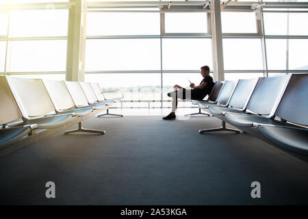 Portrait de jeune homme séduisant portant des vêtements de style décontracté assis sur le banc dans l'aéroport moderne à l'aide de terminal smartphone. Meilleur faire appel, moi Banque D'Images