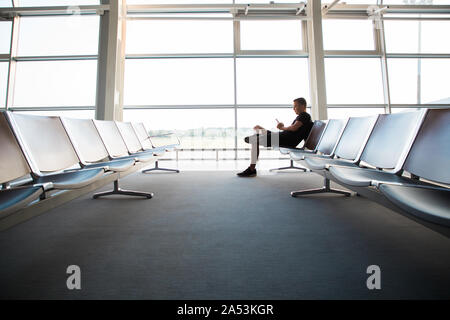 Portrait de jeune homme séduisant portant des vêtements de style décontracté assis sur le banc dans l'aéroport moderne à l'aide de terminal smartphone. Meilleur faire appel, moi Banque D'Images