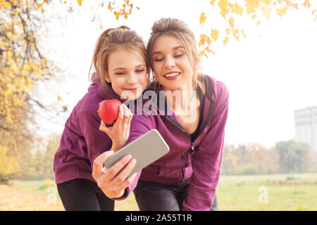 Loisirs de plein air. Soeurs article hugging in l'automne park en tenant sur smartphone selfies avec apple smiling smiling Banque D'Images