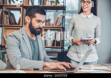 Startupers travailler ensemble au patron du bureau de travail assis sur un ordinateur portable tout en concentré femme marche à lui heureux Banque D'Images