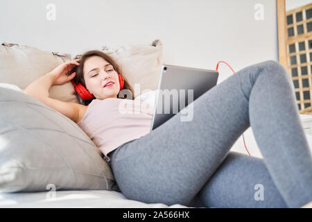 Image de jeune brunette woman with tablet dans ses mains à l'écoute de la musique tout en se trouvant sur lit dans la chambre Banque D'Images