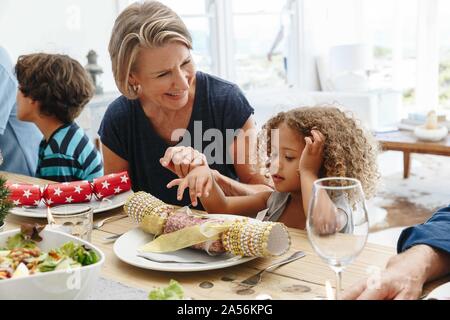 Femme parlant à petite-fille jouant avec Christmas Cracker à table à manger Banque D'Images