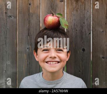 Boy smiling tout en équilibrant une pomme sur sa tête contre une clôture en bois Banque D'Images