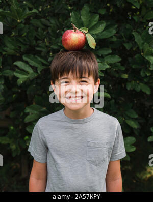 Happy Young boy équilibrer une pomme sur la tête à l'extérieur par un arbre. Banque D'Images