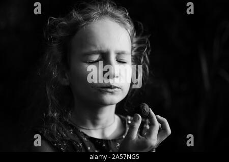 Close up portrait noir et blanc de la petite fille blonde avec queue de manger un bonbon de chocolat aux yeux clos en plein air Banque D'Images