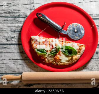 Pizza italienne typique réduit de moitié avec la roue dans une plaque rouge avec un rouleau à pâtisserie en bois à côté de lui Banque D'Images