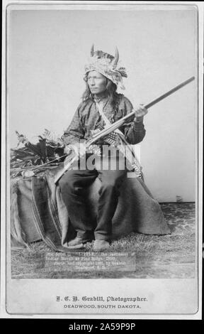 Peu d'Oglala, leader du groupe, portrait studio, assis, face vers la gauche, portant une coiffe de l'avertisseur sonore et holding rifle. Banque D'Images