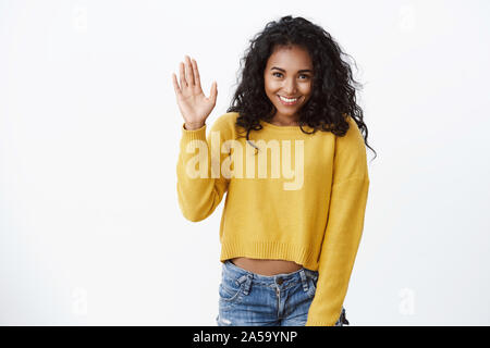 Belle timide jeune fille afro-américaine en pull jaune, curly hairstyle, se baissant comme raising hand, agitant bonjour, bonjour le geste, sourire, femme modeste Banque D'Images