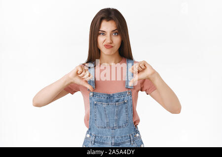 Déçu arrogant sceptique jolie femme en salopette, t-shirt, un sourire satisfait, showing thumbs-vers le bas et de froncer en désaccord, express Banque D'Images