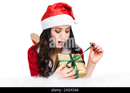 Jeune femme enthousiaste de l'ouverture d'un cadeau de Noël. Jolie fille dans un costume de Père Noël déclenche un ruban sur un cadeau de nouvel an allongé sur le lit sur isolé sur Banque D'Images