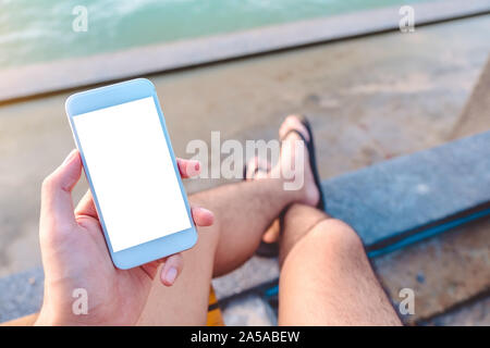 Image d'une maquette man's hand holding white téléphone mobile avec écran de bureau vierge assis au bord de la mer Banque D'Images