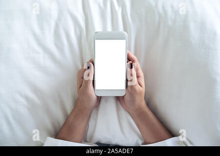 Vue de dessus de l'image immersive a woman's hand holding white téléphone mobile avec écran de bureau vide sur l'oreiller et assis dans le lit Banque D'Images