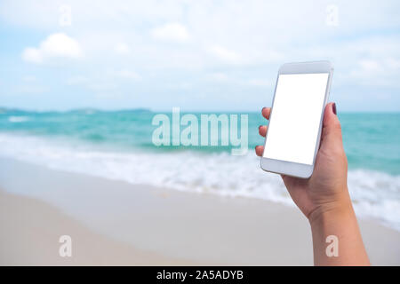Image maquette de woman's hand holding white téléphone mobile avec écran de bureau vide en face de la mer et fond de ciel bleu Banque D'Images