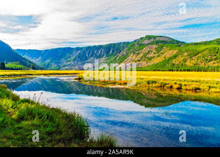 La vallée de la rivière qui traverse les champs d'herbe jaune doré avec ses montagnes couvertes de forêts sous ciel nuageux avec un bleu lumineux. En raison de l'eau. Banque D'Images