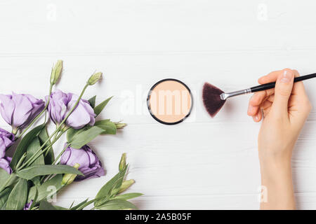 Femme main tenant un miroir à l'aide pinceau poudre compacte sur fond blanc avec des fleurs lisianthus, télévision jeter la composition. Banque D'Images