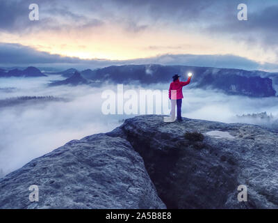 Fille de tourisme traveler se dresse au sommet de la montagne jusqu'à une lumière forte holding sa tête et des éclairs à l'obscurité. Femme conserve l'ampoule flash dans les mains dans misty da Banque D'Images