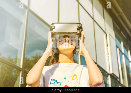 Belle jeune femme en blanc T-shirt à l'aide de casque de réalité virtuelle maintenant ses mains sur les côtés et souriant. Bâtiment moderne en verre entreprise facad Banque D'Images