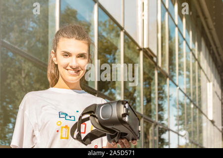 Belle jeune femme en blanc T-shirt holding casque de réalité virtuelle et regardant droit dans la caméra. Entreprise moderne en verre en façade du bâtiment Banque D'Images